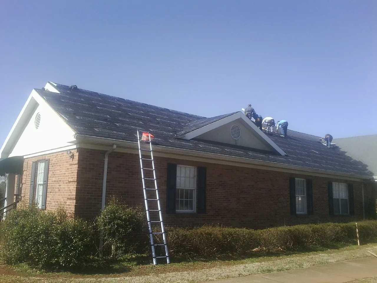 Crew tearing shingles off a brick ranch roof with a ladder set — Metal Roof Repair work by our crew in Port Crane