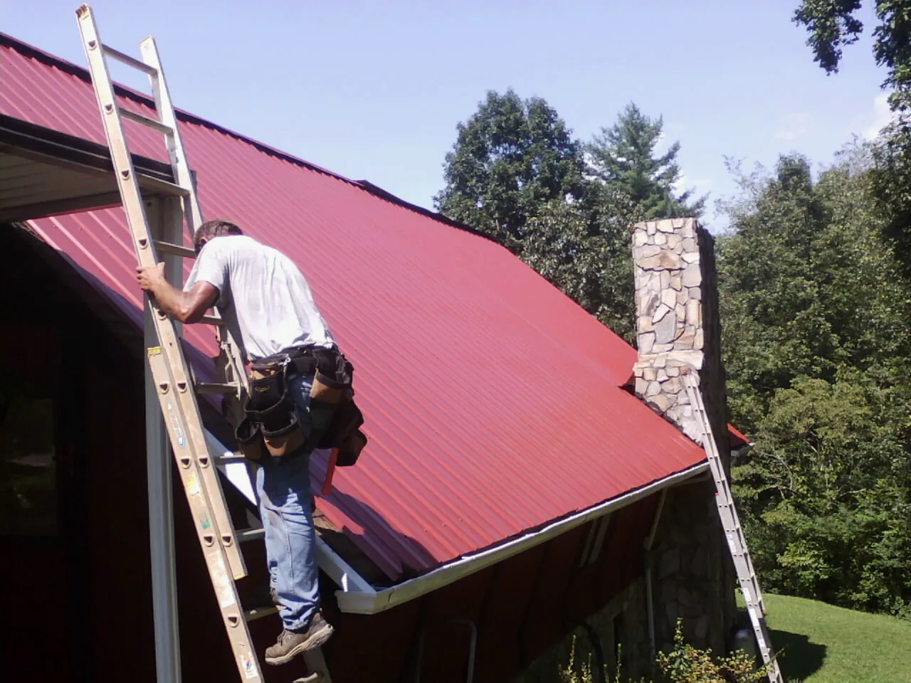 Roofer on a ladder beside a red metal roof and stone chimney — Soffit & Fascia Repair by Roofing Brothers in Port Crane